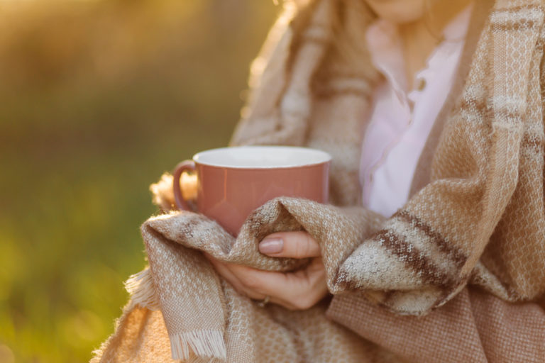 Inverno ayurvédico: mulher enrolada em um cobertor xadrewz em tons de bege segurando uma xícara de chá ao por do sol