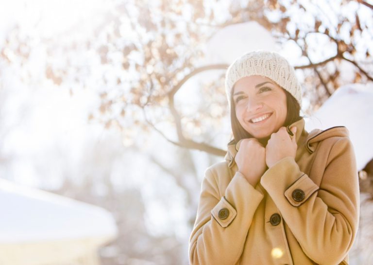 inverno ayurvédico: Mulher sorridente ao ar livre em um ambiente iluminado, porém coberto de neve. Ela está vestida com um casaco marrom claro e usando uma touca branca.