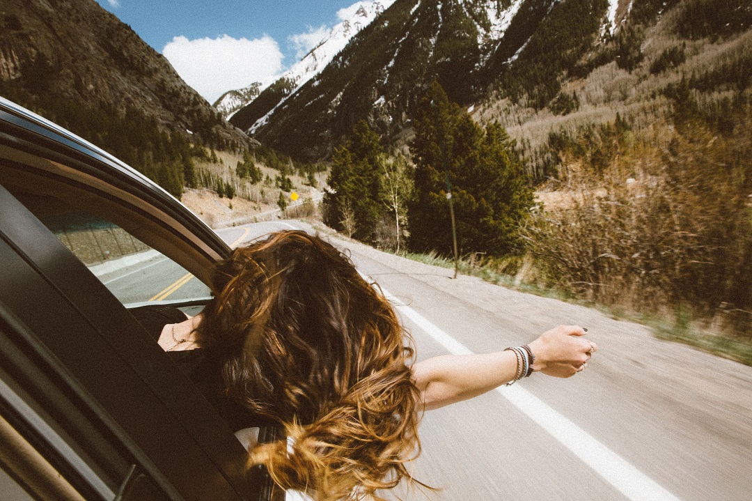 equilíbrio ayurvédico: mulher com parte do corpo para fora do carro, o braço estendido, enquanto o carro está seguindo em uma estrada em meio as montanhas. Ela está de blusa preta e tem os cabelos claros