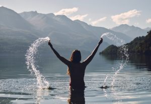 Maha Gunas: mulher em um lago, de costas ao por do sol levntando água coma s mãos em uma paisagem montanhosa