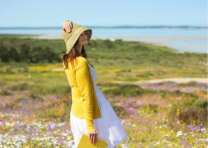 Primavera: mulher de vestido branco e casaco amarelo usando um chapéu de palha parada em um campo florido em frente a uma imensa lagoa